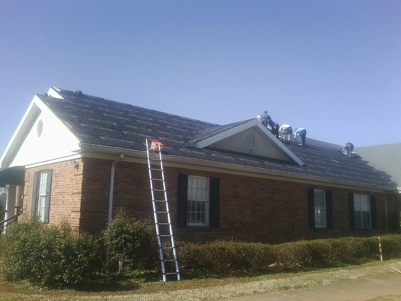 Crew tearing shingles off a brick ranch roof with a ladder set — Emergency Roof Repair work by our crew in Postville