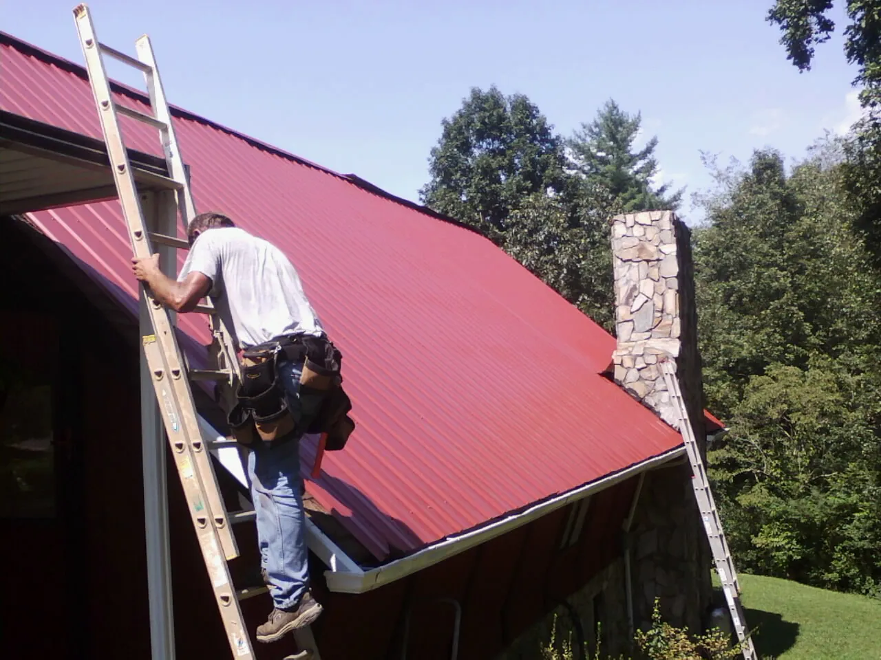 Roofer on a ladder beside a red metal roof and stone chimney — Soffit & Fascia Repair by Roofing Brothers in Postville